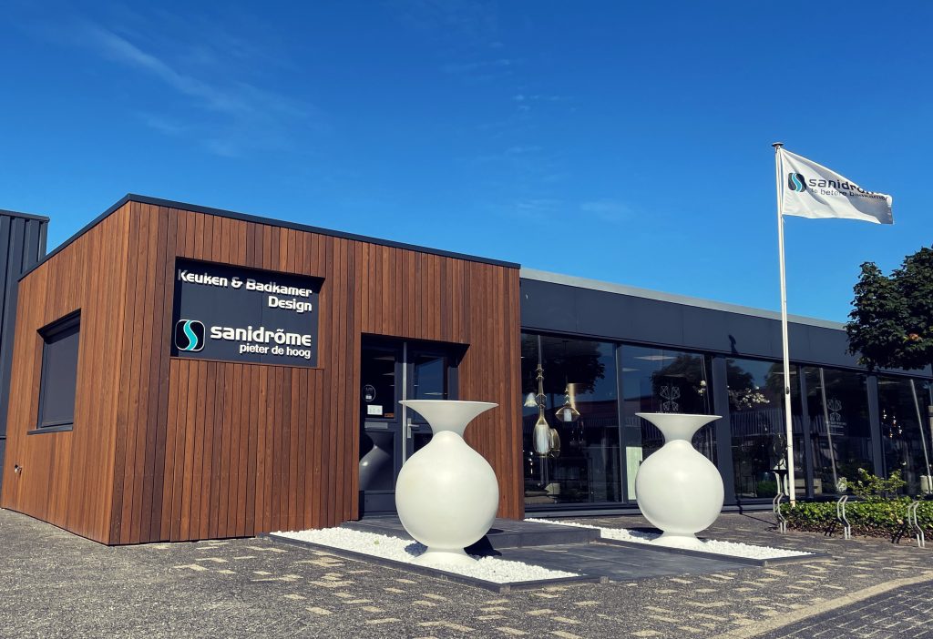 Vooraanzicht van de showroom van Sanidrõme Pieter de Hoog met moderne houten gevel, grote design potten en Sanidrõme-vlag bij helderblauwe lucht.