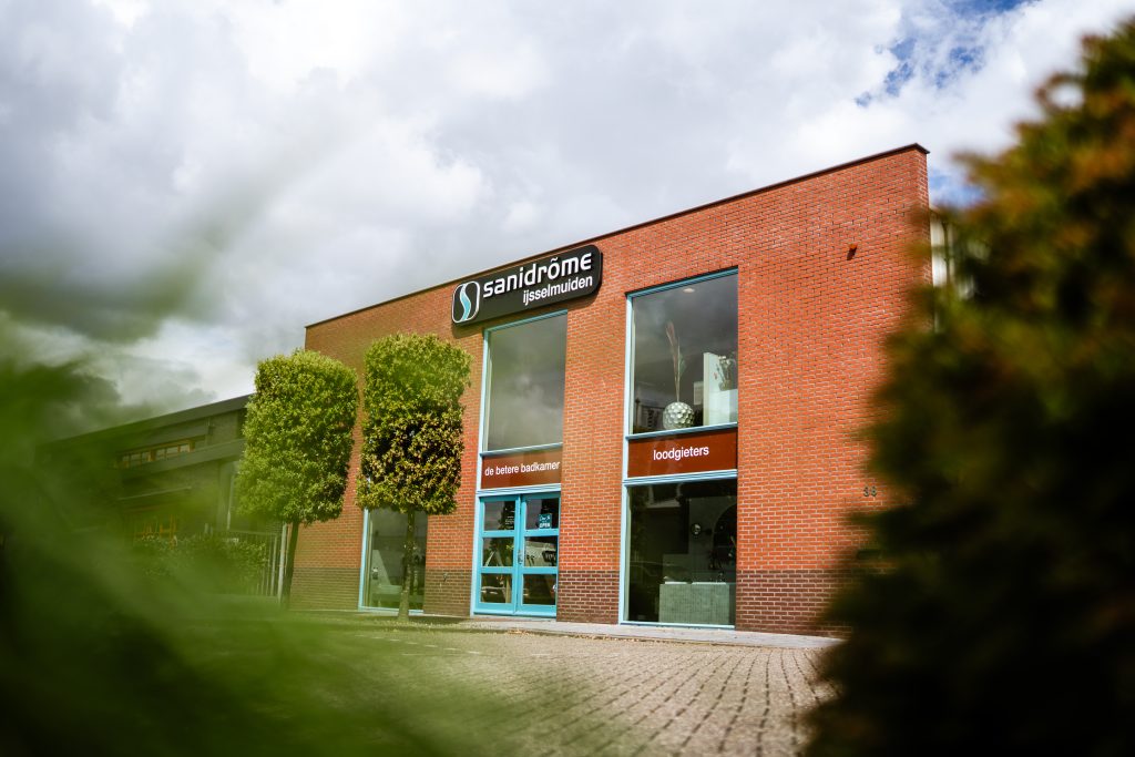 Vooraanzicht van Sanidrõme IJsselmuiden met showroomgevel en opvallend logo op rode bakstenen gevel.